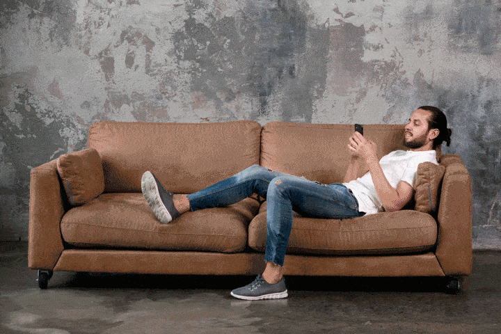 A man in casual clothes lounges on a brown sofa, using his smartphone and smiling, with one leg stretched out and the other bent. In the backdrop of a textured gray wall, he embodies how made-to-measure businesses can still thrive in a post-COVID world.
