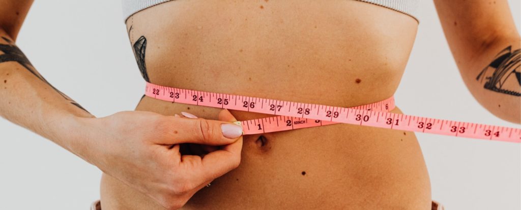A woman is taking body measurements at home with a pink measuring tape.