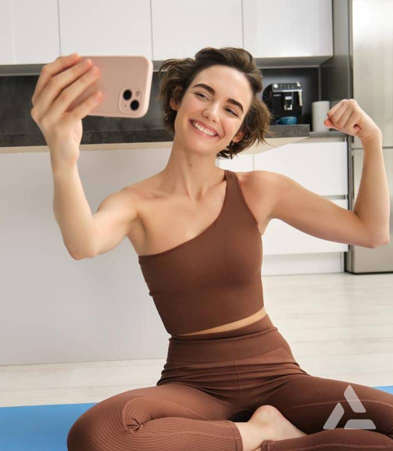 A woman in brown athletic wear, sitting on a yoga mat, takes a selfie while flexing her arm and smiling during her AI-powered body scanning session.