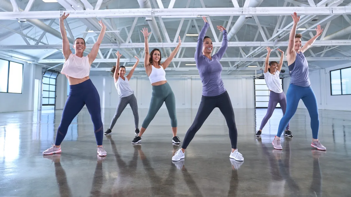 A group of six women in sportswear joyfully performing aerobics in a spacious, brightly lit studio, equipped with AI-powered body scanning technology.