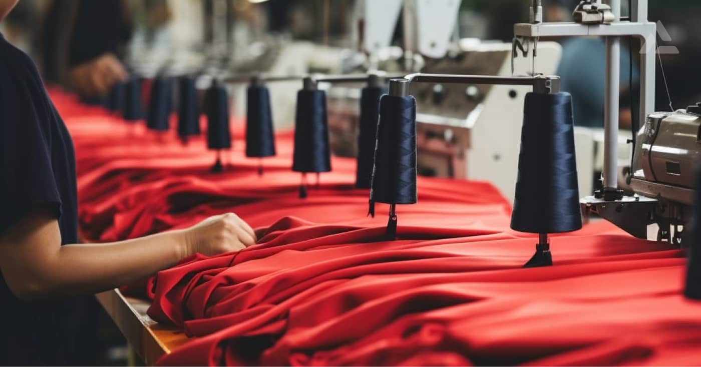 A worker cuts red fabric at a table with several reels of thread and sewing machines in a textile factory specializing in on-demand fashion.
