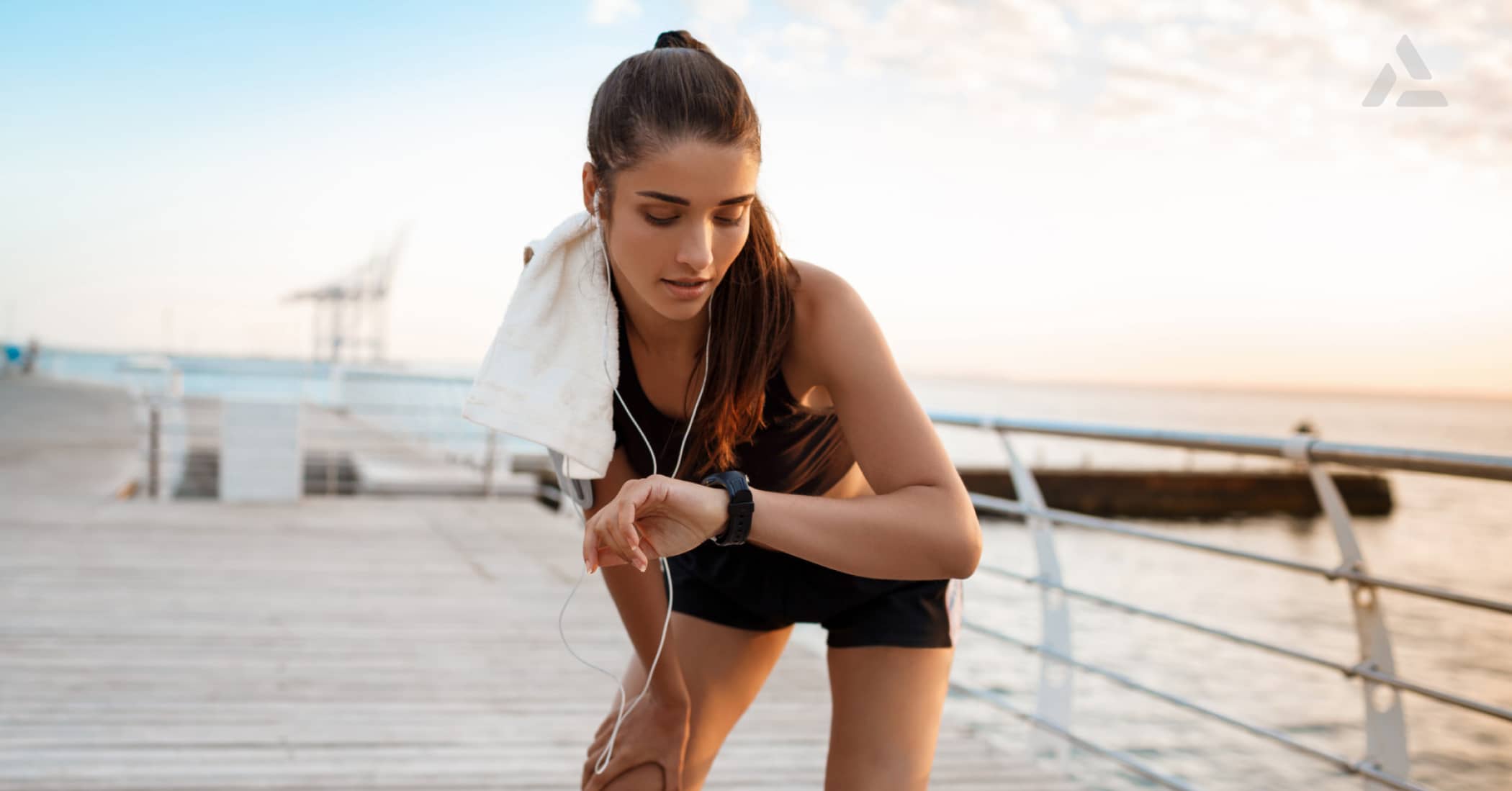 A woman in workout attire with a towel around her neck looks at her smartwatch on a boardwalk by the sea during sunrise or sunset, embodying the future of connected fitness as highlighted in the 2024 Guide.