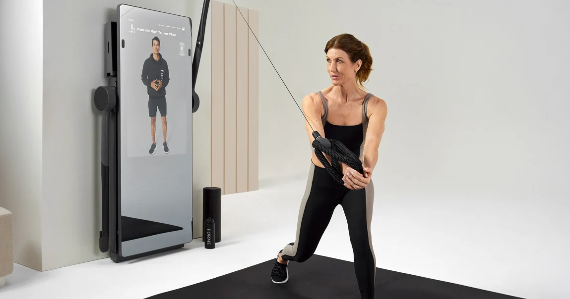 A woman performs a workout using a cutting-edge fitness machine with a digital trainer screen in a minimalistic room, showcasing the latest innovation from top fitness tech companies in 2024.