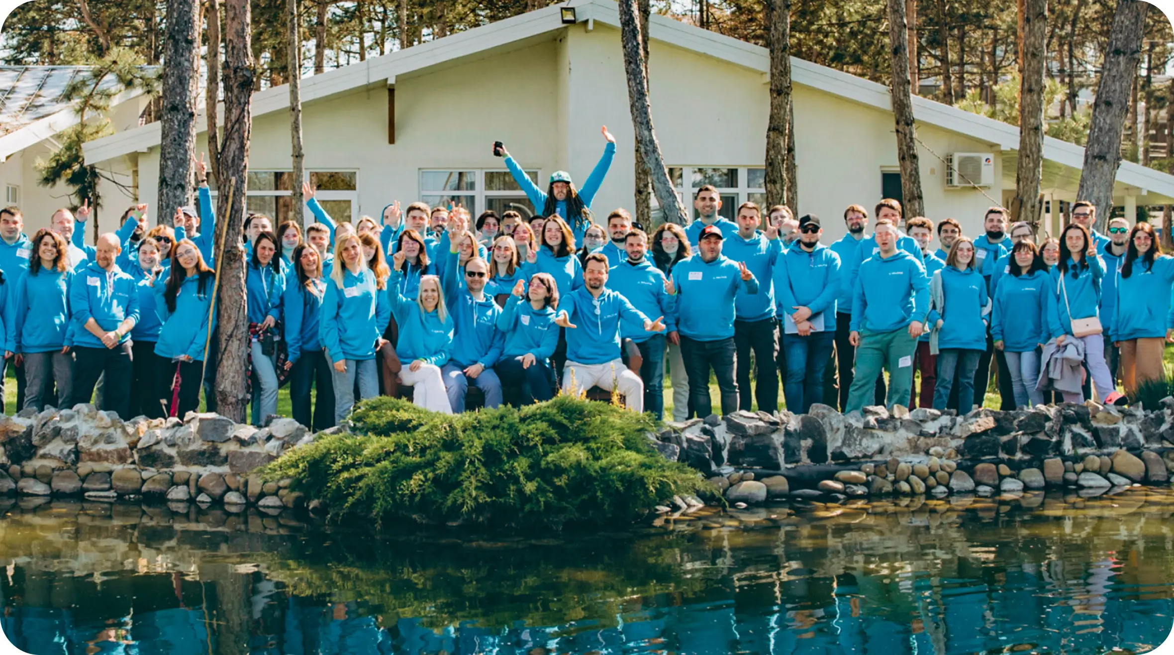 Group of people in blue shirts posing cheerfully by a pond in front of cabins at a daytime outdoor gathering, "New About Us" featured prominently on their attire.