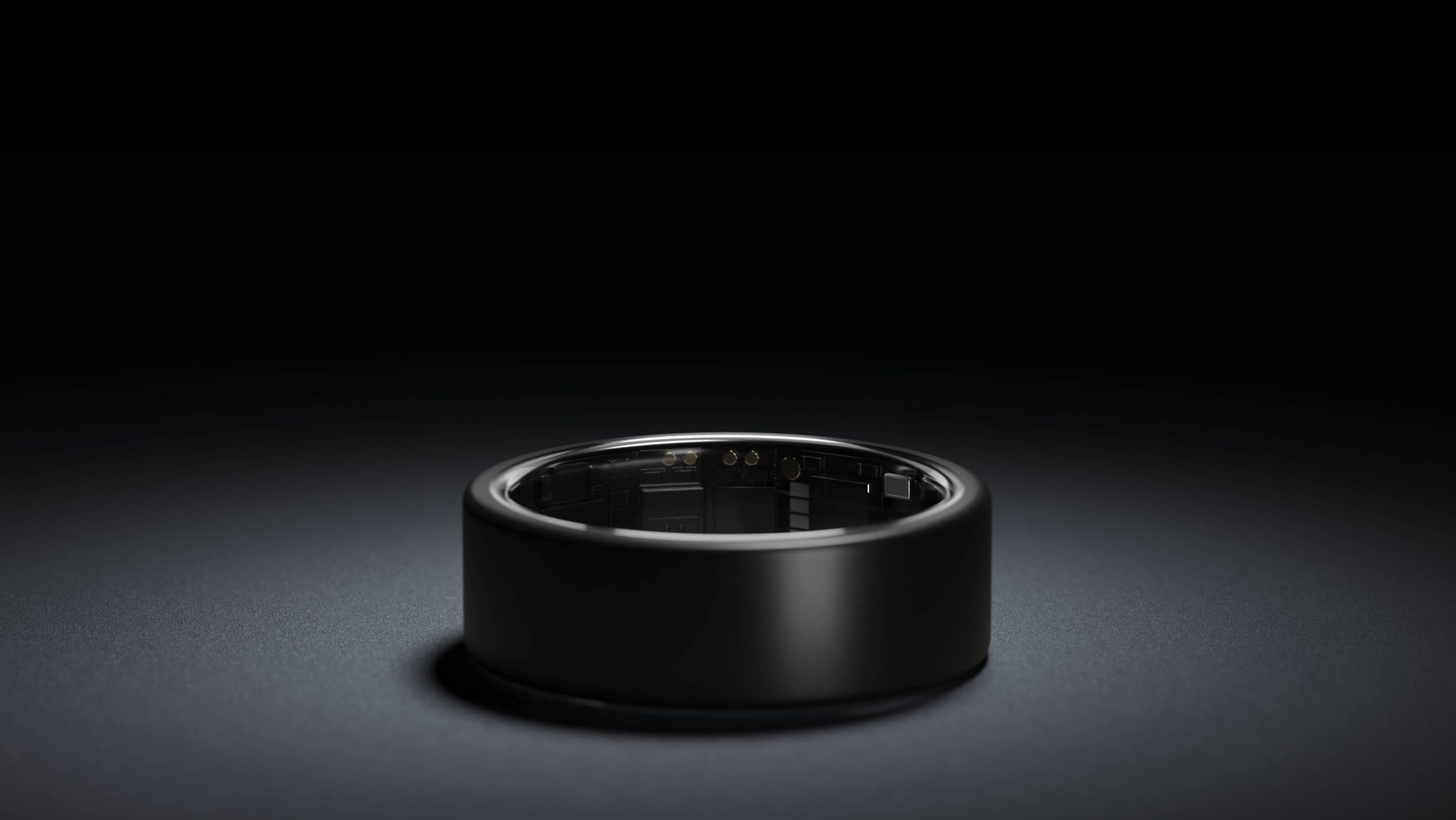 A black smart ring with a sleek design is placed on a dark surface, illuminated by soft lighting. The interior of the ring reveals technical components, showcasing its role in the connected fitness industry.