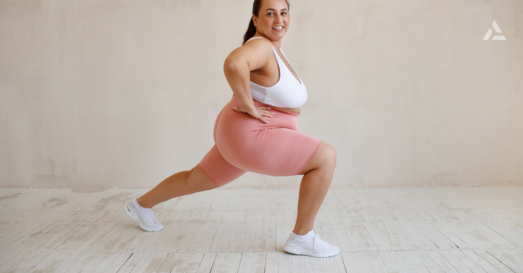 A woman in a white sports bra and pink shorts is doing a forward lunge exercise indoors on a light wooden floor, demonstrating all you need to know for effective weight loss.