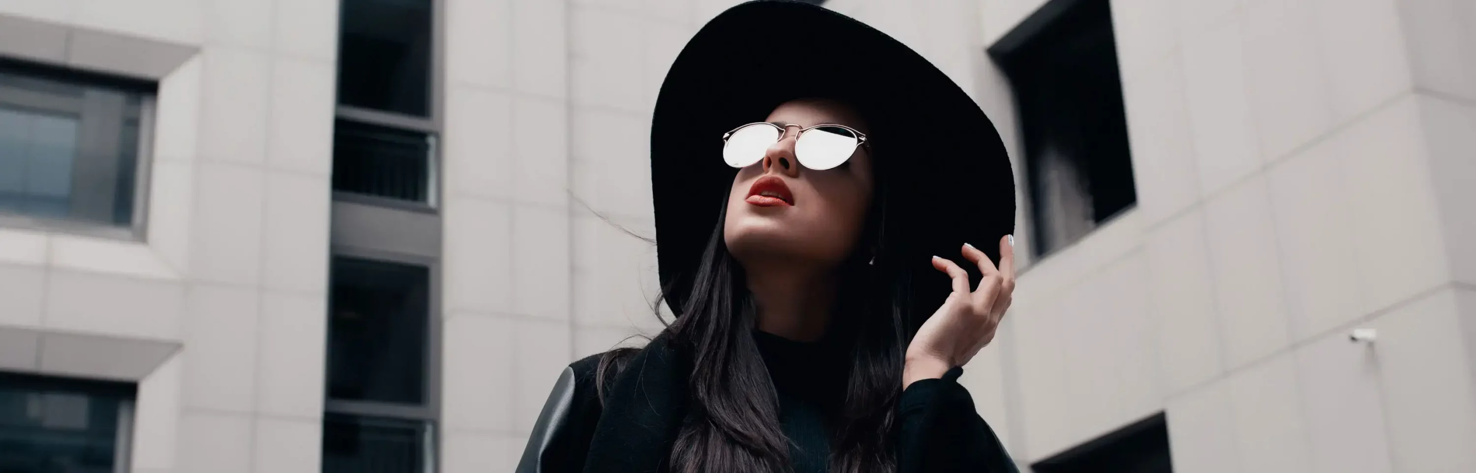 A person wearing a black hat and mirrored sunglasses looks up while standing in an urban area with modern buildings in the background, showcasing a sleek, redesigned look for YourFit 2024.
