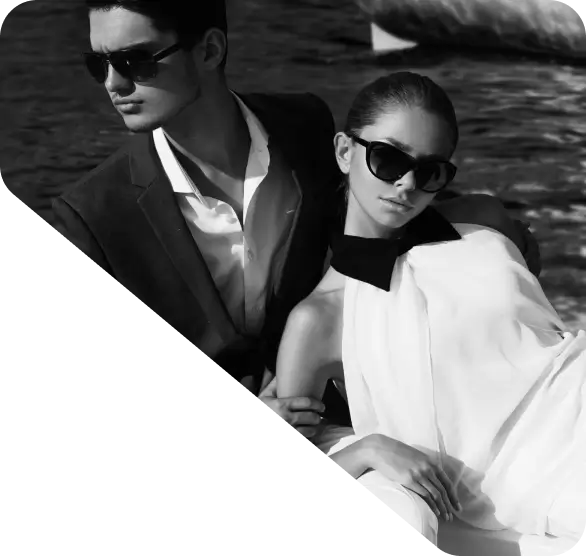 A man and a woman wearing sunglasses lean against each other in a black-and-white photo with a boat on water in the background, perfect for your homepage.