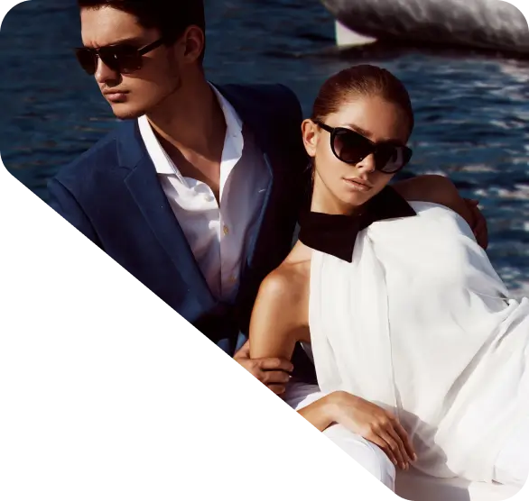 A man and woman dressed elegantly in sunglasses pose near water. The important detail to note is the man's blue suit and the woman's white dress with a black collar, perfect for any homepage feature.