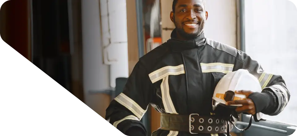 A firefighter wearing a black uniform with reflective stripes smiles and holds a white helmet inside a building, perfectly capturing the essence for our website's homepage.