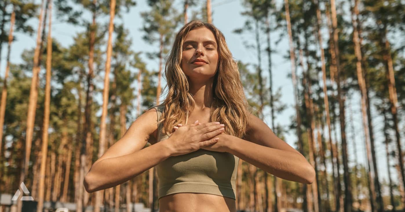 A woman with long hair stands outdoors with tall trees in the background, holding her hands to her chest and eyes closed, appearing to be practicing mindfulness or meditation, embodying one of the top fitness trends for 2024.