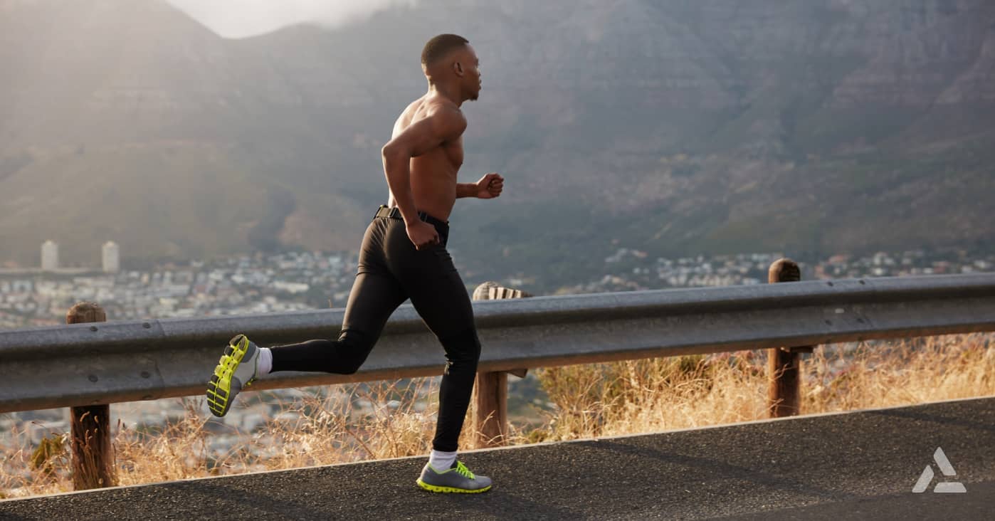 A man runs on a paved road next to a metal guardrail, with a cityscape and mountains in the background, embodying 2024 fitness industry trends.