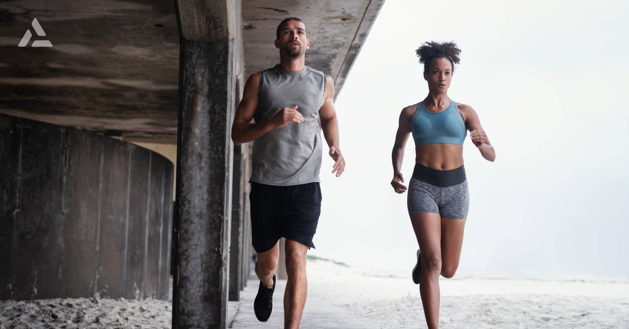 A man and a woman jog side-by-side under a concrete structure on the beach, embodying the latest fitness industry trends of 2024.