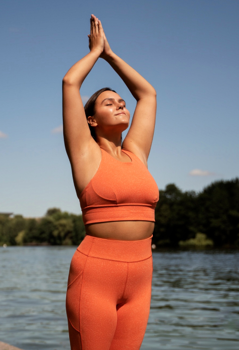 A person in orange activewear gracefully practices yoga by a tranquil lake under the clear blue sky, framed by redesigned trees in the background, capturing a moment that feels straight out of an inspiring 2024 ebook.