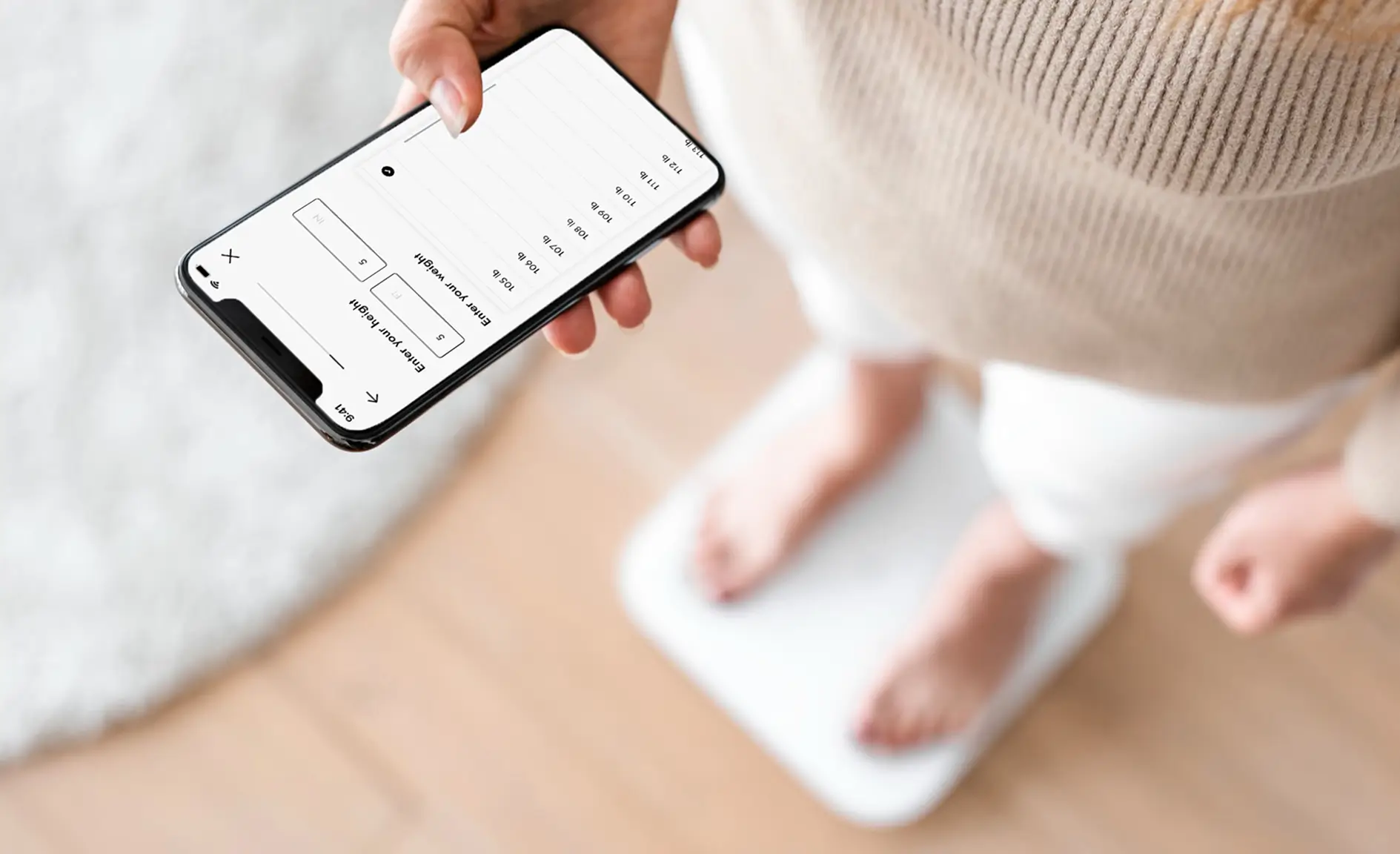 A person stands on a digital scale for BMI verification, holding a smartphone that displays a weight tracking app.