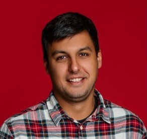 A man with short dark hair wearing a red, white, and black plaid shirt stands in front of a solid red background, smiling slightly at the camera.